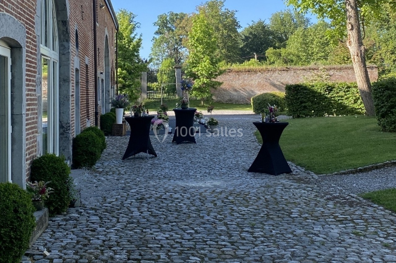 Allée pavée bordée de verdure avec des tables hautes noires décorées de fleurs, devant un bâtiment en briques.