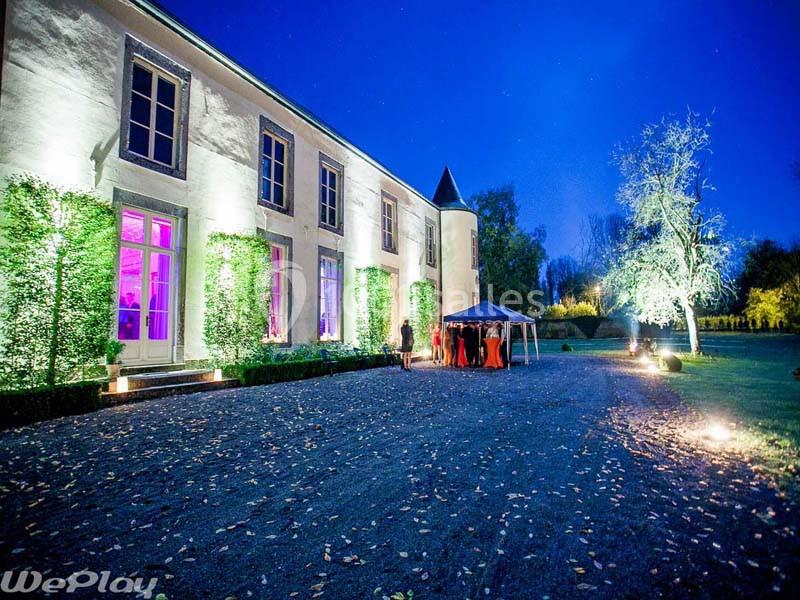 Façade éclairée d'un château entouré de végétation, avec des invités rassemblés sous une tente le soir.