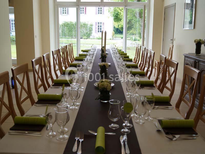 Table dressée avec des chaises en bois, nappes blanches, chemins de table marron, serviettes vertes et vue sur un jardin.