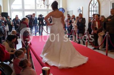 Une femme en robe de mariée défile sur un tapis rouge devant un public dans une salle éclairée par des lustres.