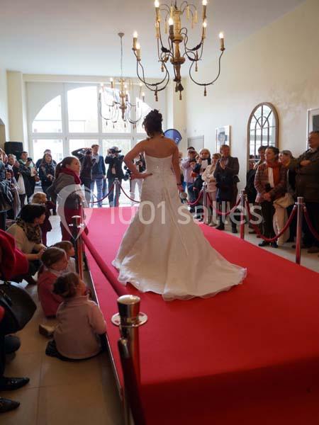 Une femme en robe de mariée défile sur un tapis rouge devant un public dans une salle éclairée par des lustres.