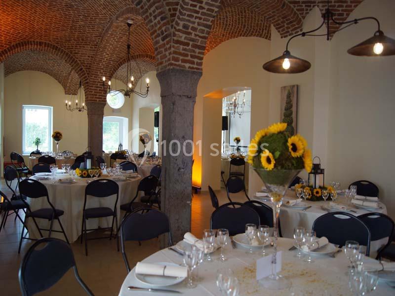 Salle de réception avec tables rondes dressées, décorées de tournesols, sous des voûtes en briques apparentes.