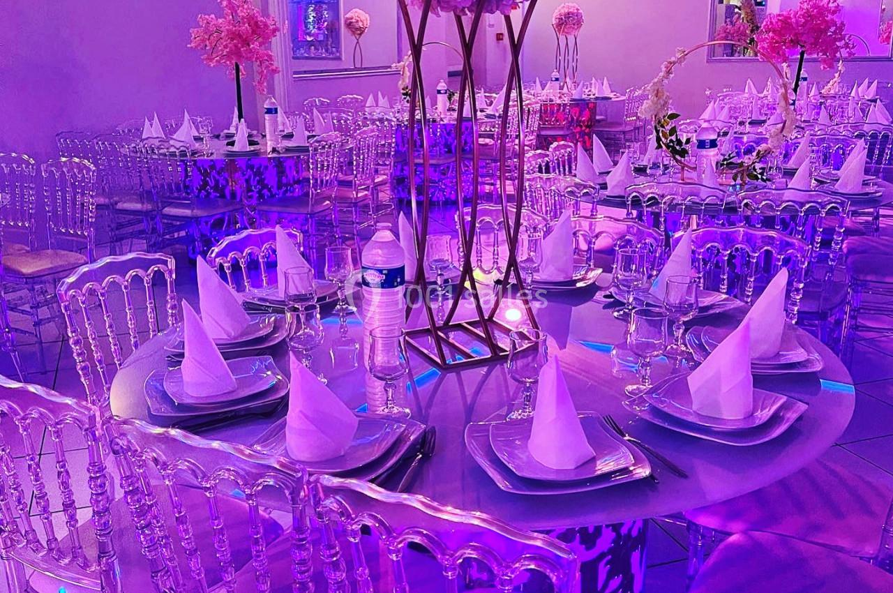 Salle de réception décorée avec des tables rondes, nappes violettes, chaises transparentes et centre de table floral.