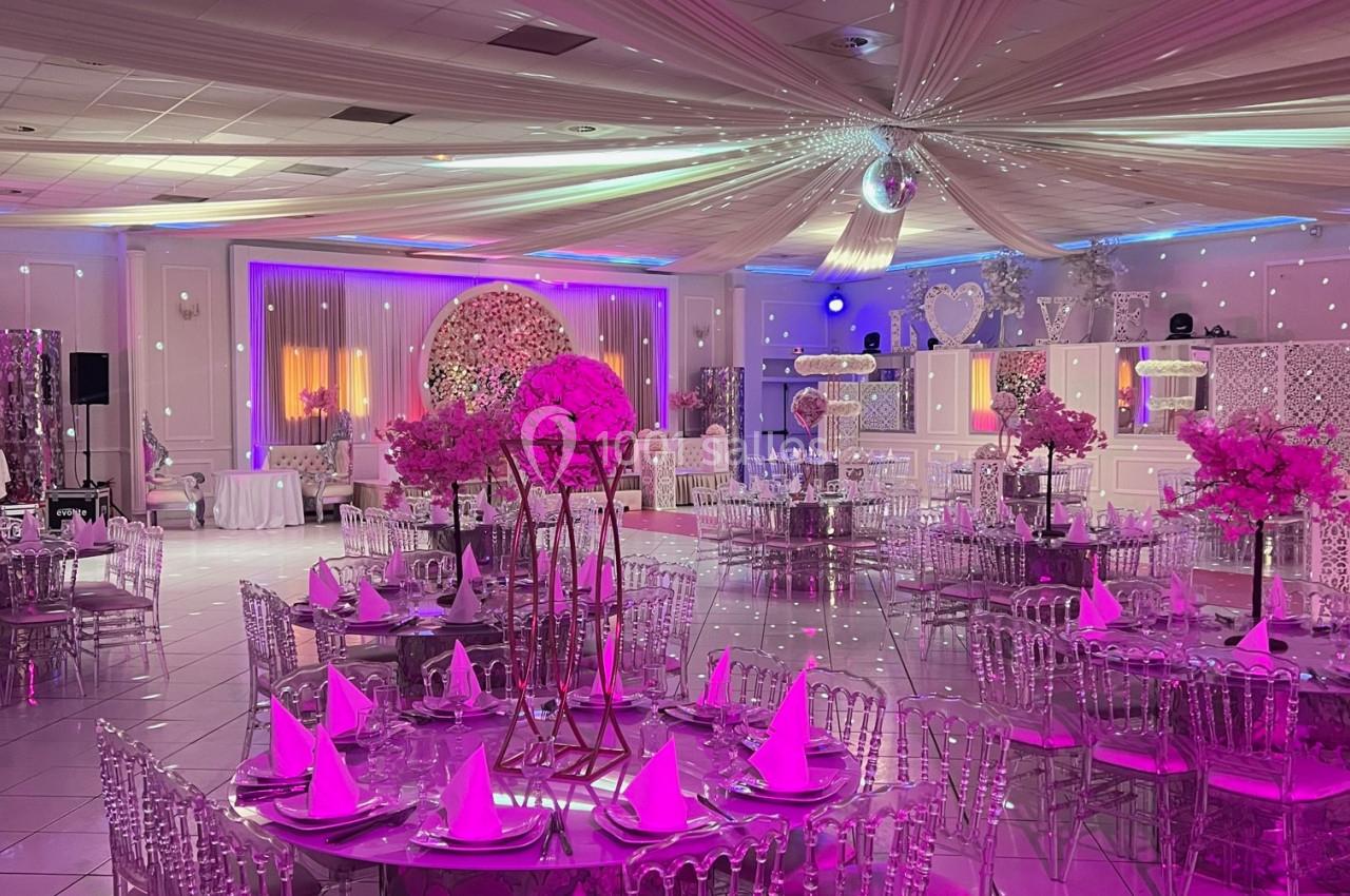 Salle de réception décorée avec des tables rondes, nappes roses, chaises transparentes et éclairage tamisé rose.