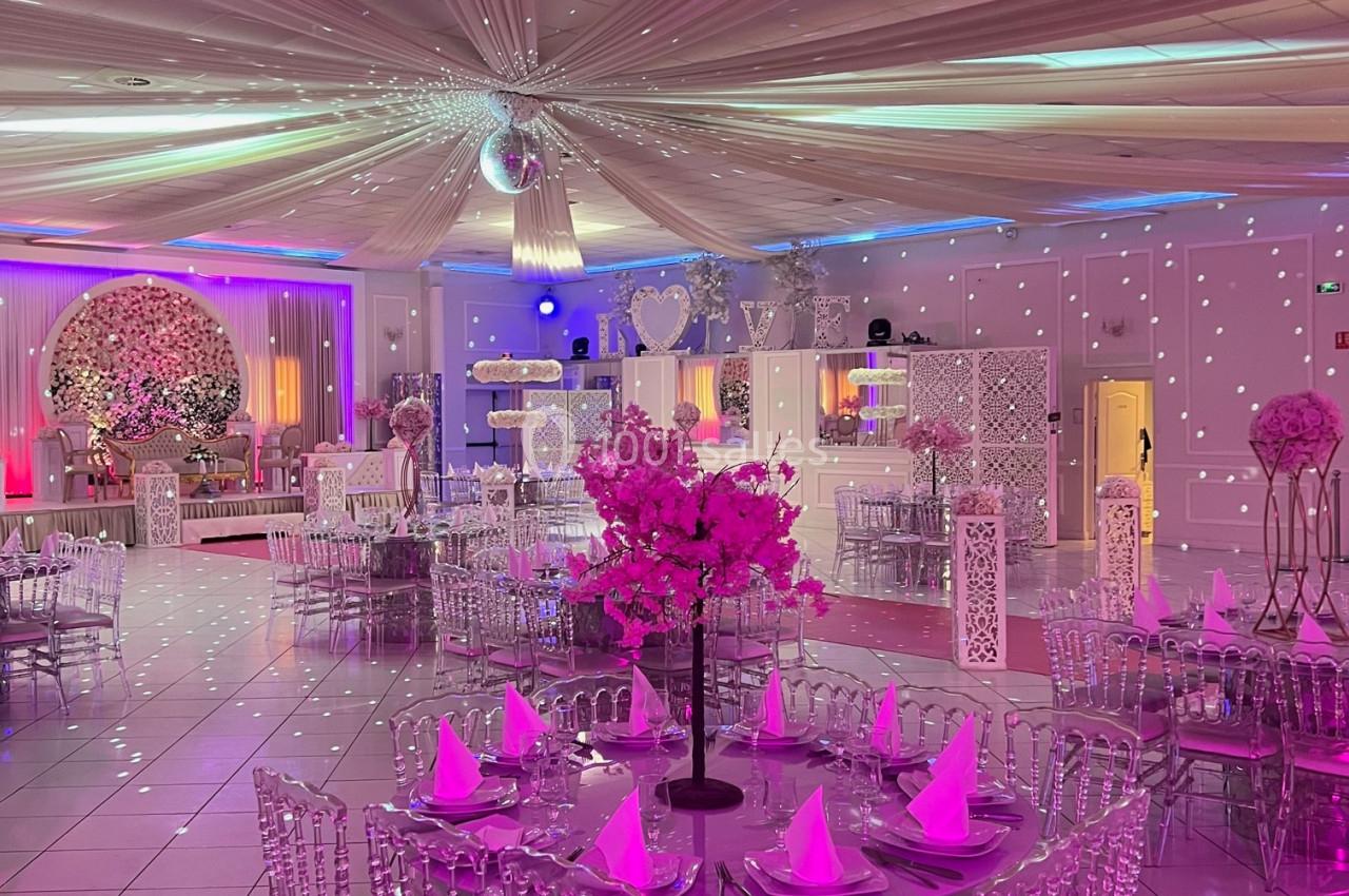 Salle de réception décorée avec des tables rondes, chaises transparentes et éclairage coloré créant une ambiance festive.