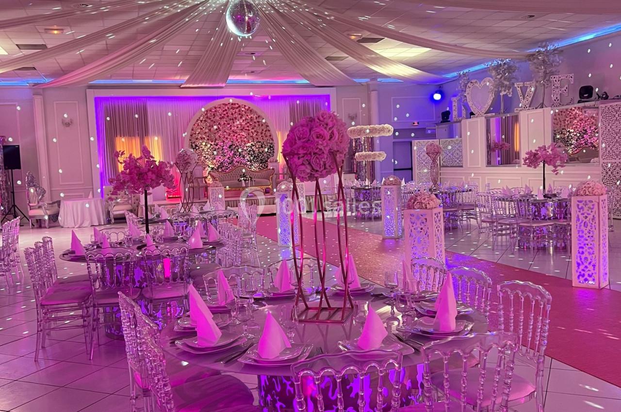 Salle de réception décorée avec des tables rondes, chaises transparentes et éclairage rose et violet.