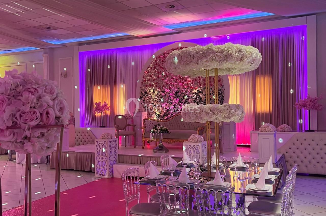 Salle de réception décorée avec des tons roses et violets, tables dressées et mur floral illuminé en arrière-plan.