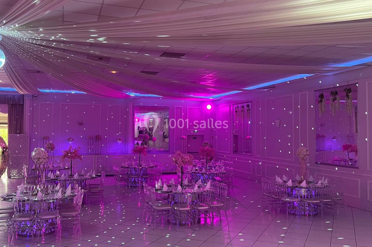 Salle de réception décorée avec des tables dressées, éclairage rose et plafond orné de drapés blancs.