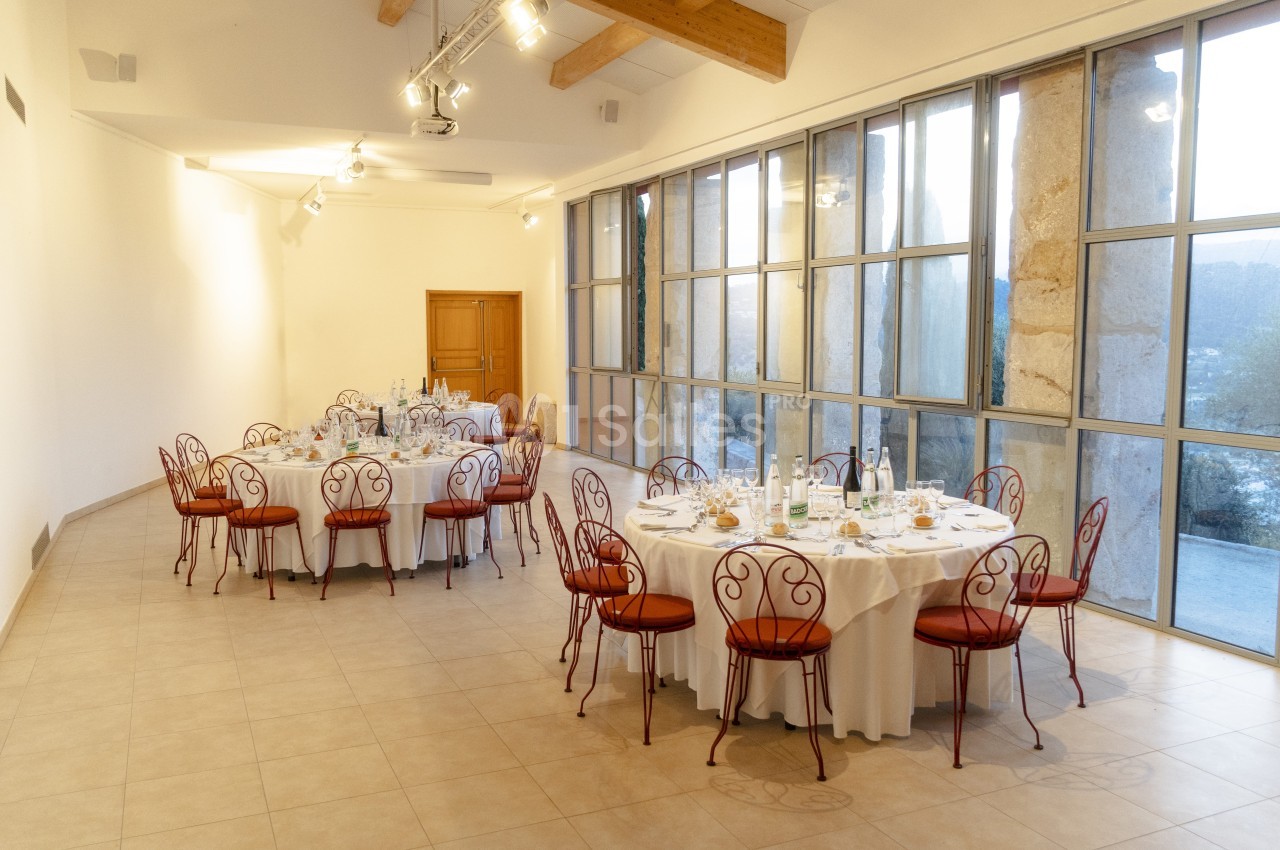 Salle lumineuse avec tables rondes dressées, chaises en fer forgé et grandes baies vitrées donnant sur l'extérieur.