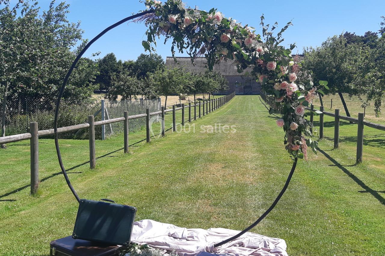 Arche circulaire décorée de fleurs roses et feuillages, installée sur une pelouse avec un chemin bordé de clôtures en bois.