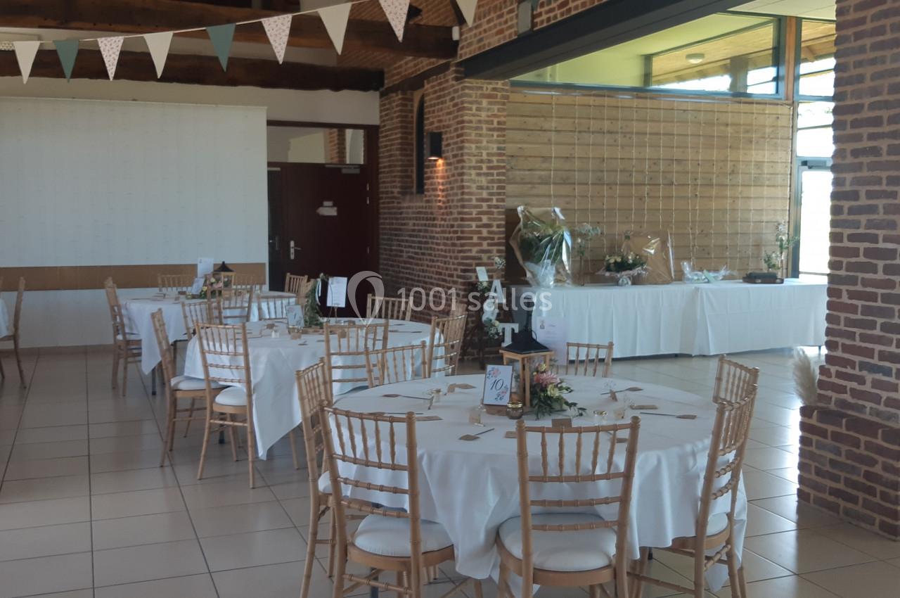 Salle de réception décorée avec des tables rondes, nappes blanches, chaises en bois clair et guirlandes suspendues.