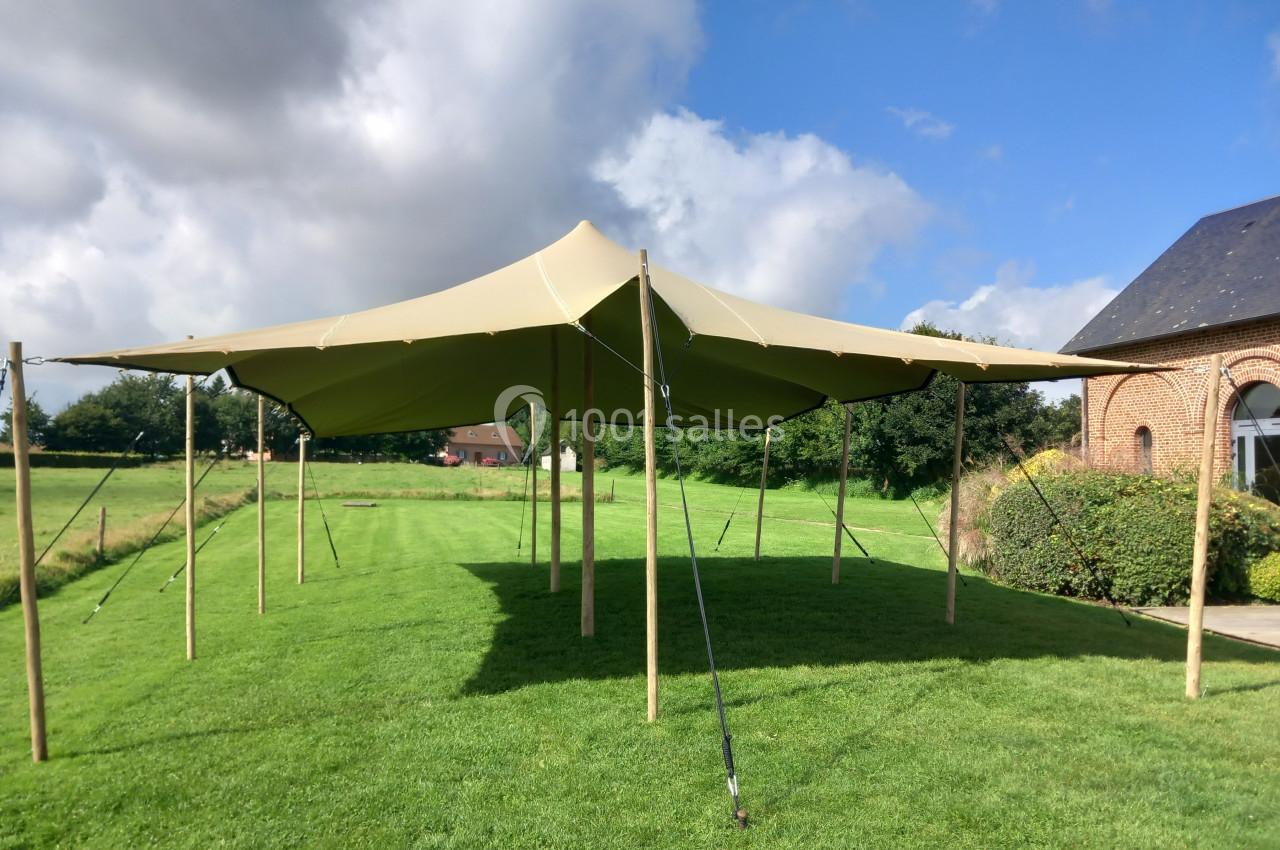 Tente beige montée sur une pelouse, soutenue par des poteaux en bois, avec un bâtiment en briques à droite.