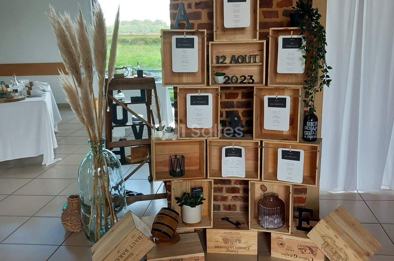 Mur décoratif en caisses en bois avec des plans de table, des éléments végétaux et des objets rustiques pour un mariage.