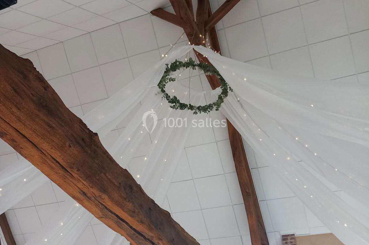 Guirlande lumineuse et couronne végétale suspendues sous une charpente en bois avec drapés blancs.