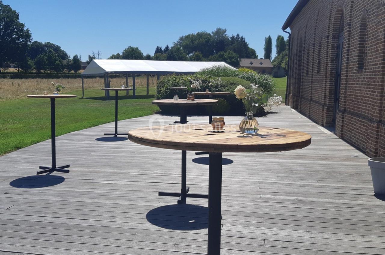 Tables rondes en bois disposées sur une terrasse en bois, avec vue sur un jardin et une tente blanche au fond.