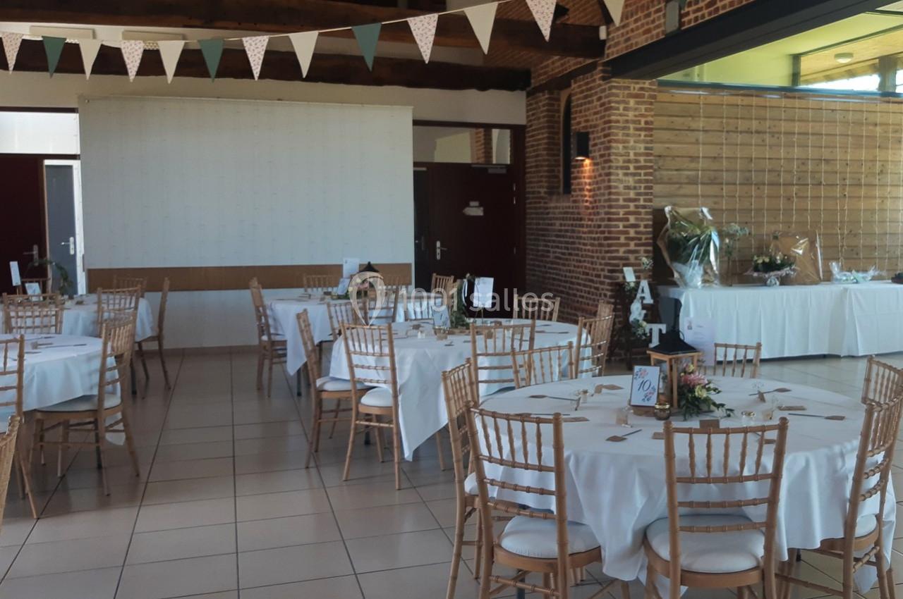 Salle décorée pour un événement avec des tables rondes dressées, des chaises en bois et des guirlandes suspendues.