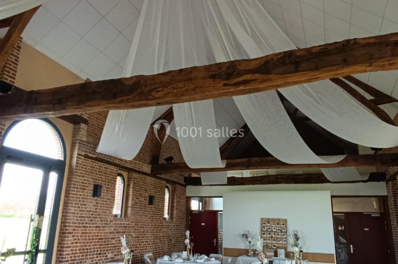 Salle de réception décorée avec des tables rondes dressées, nappes blanches et voilages suspendus au plafond.