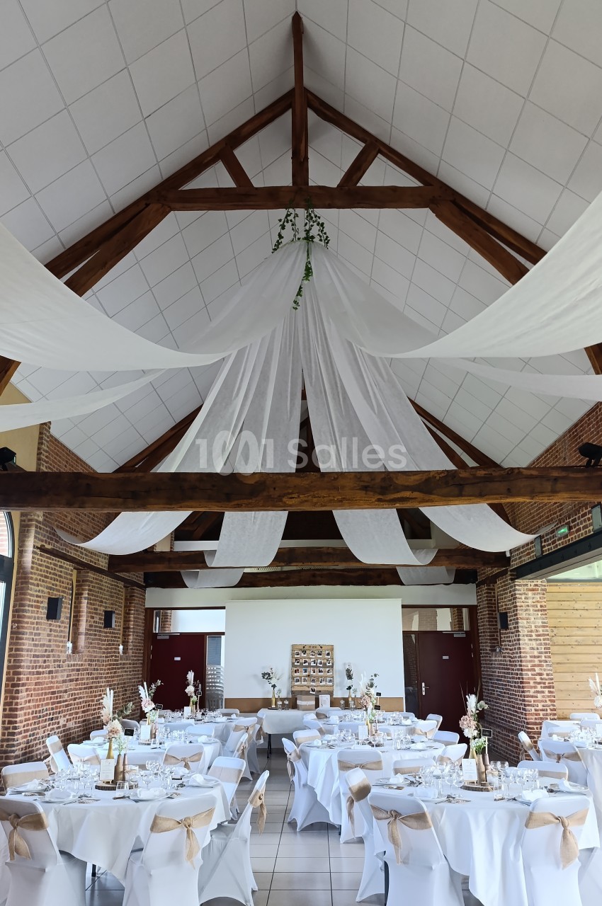 Salle de réception décorée pour un mariage, avec tables dressées, nappes blanches et voilages suspendus au plafond.