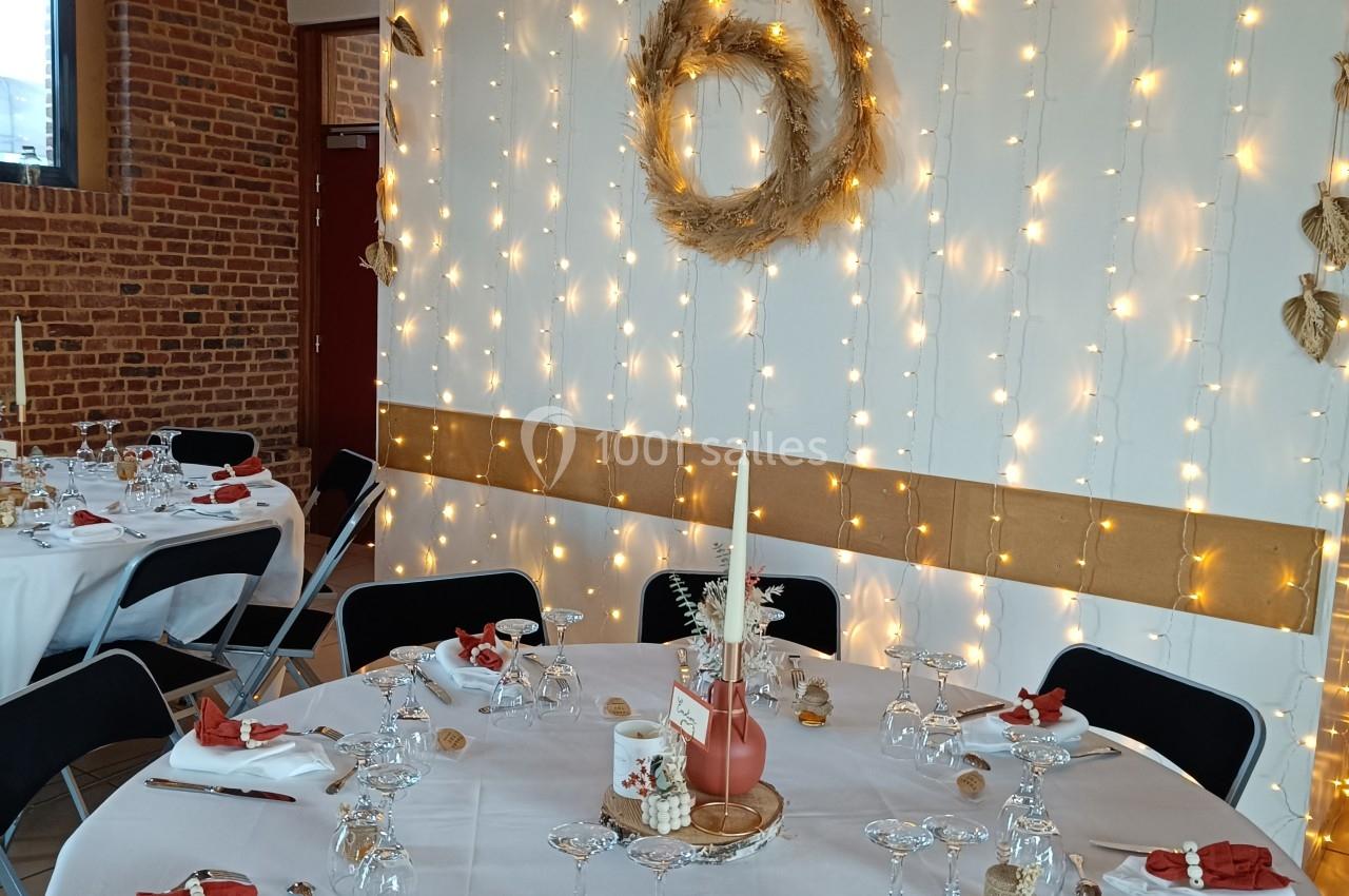 Salle décorée pour un événement avec tables dressées, nappes blanches, serviettes rouges et guirlandes lumineuses sur un mur.