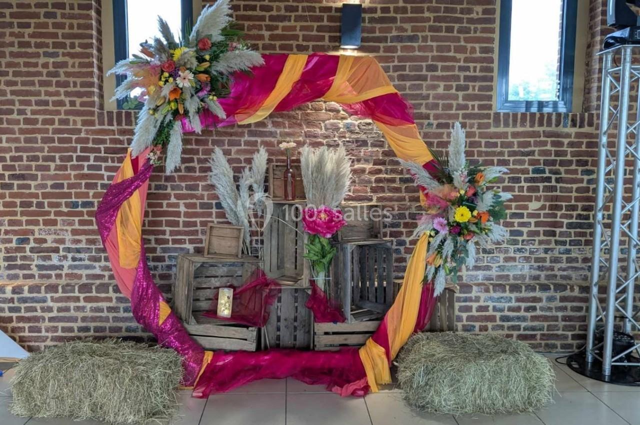 Cercle décoratif avec tissus colorés, fleurs et bottes de paille, devant un mur de briques dans un espace intérieur.