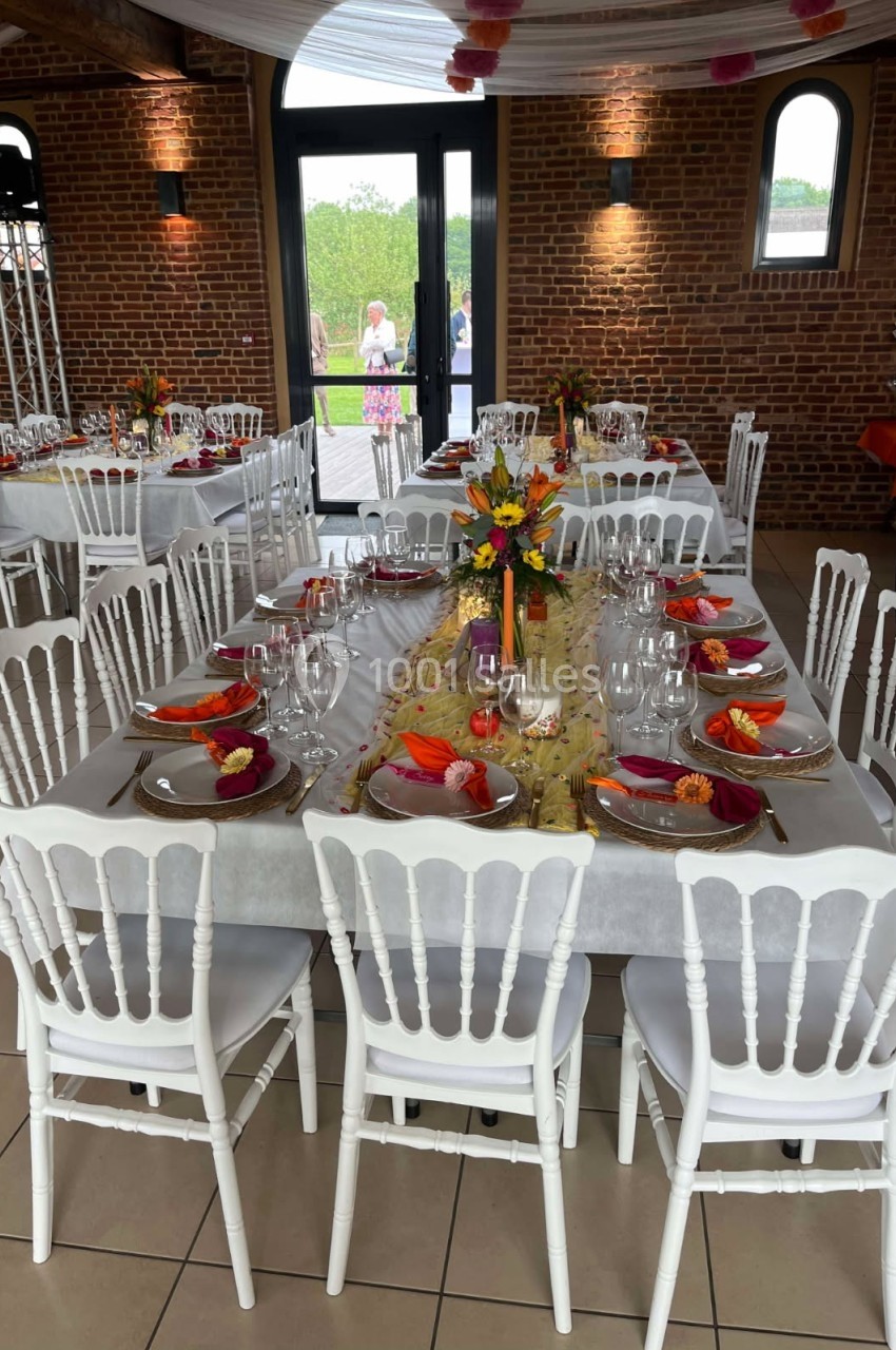 Salle décorée pour un repas festif avec tables dressées, nappes blanches, chaises blanches et compositions florales colorées.