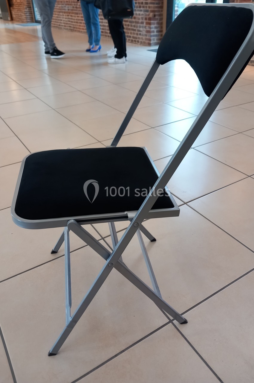 Chaise pliante noire avec structure métallique argentée, posée sur un sol carrelé beige dans un espace intérieur.