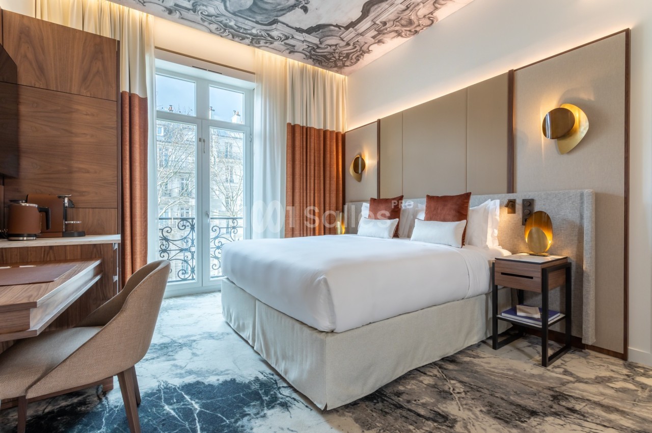Chambre d'hôtel lumineuse avec lit double, bureau en bois, fauteuil, et grande fenêtre donnant sur un balcon.