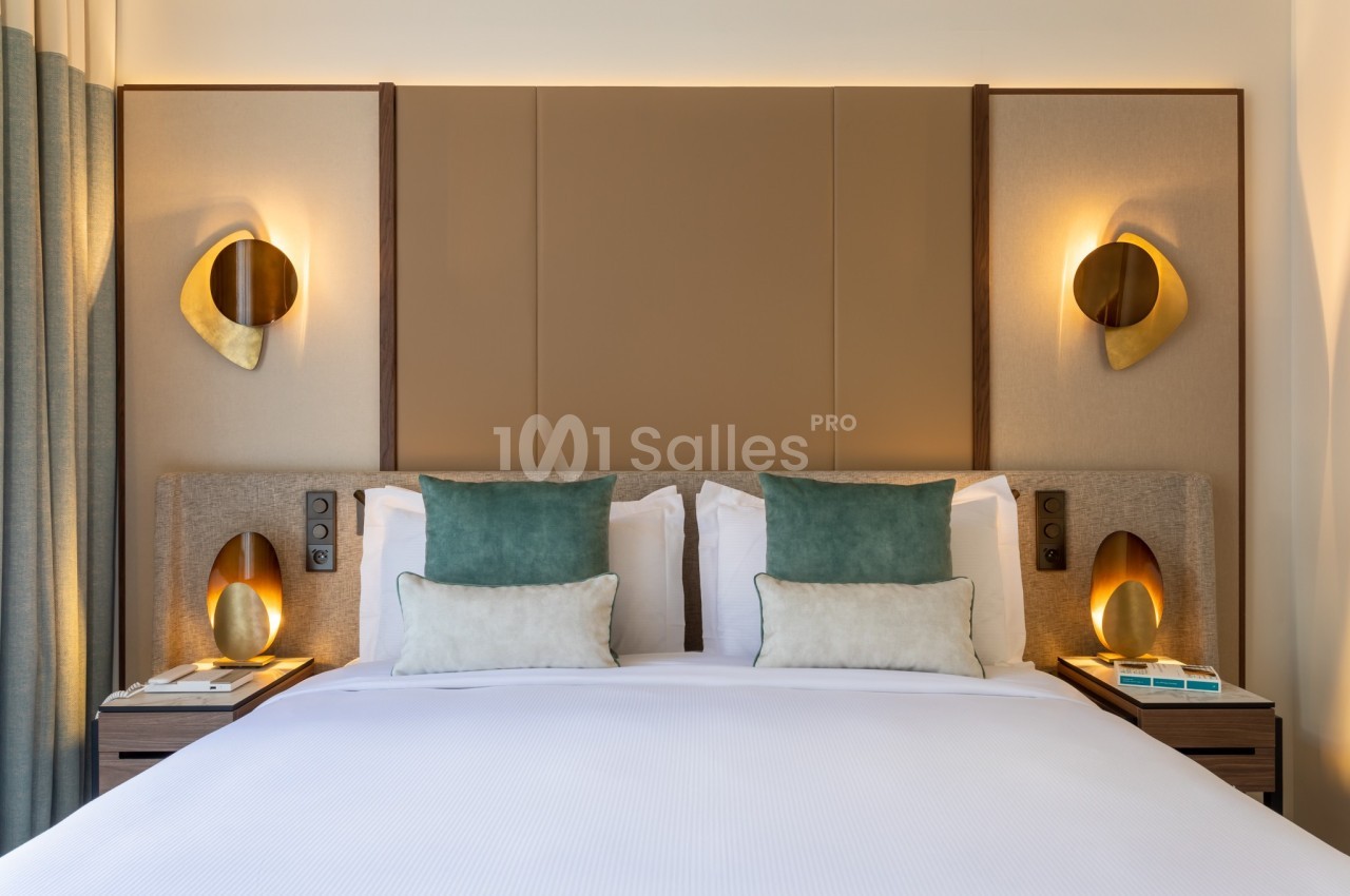 Lit double avec tête de lit rembourrée, coussins verts et beige, éclairage mural doré et tables de chevet.