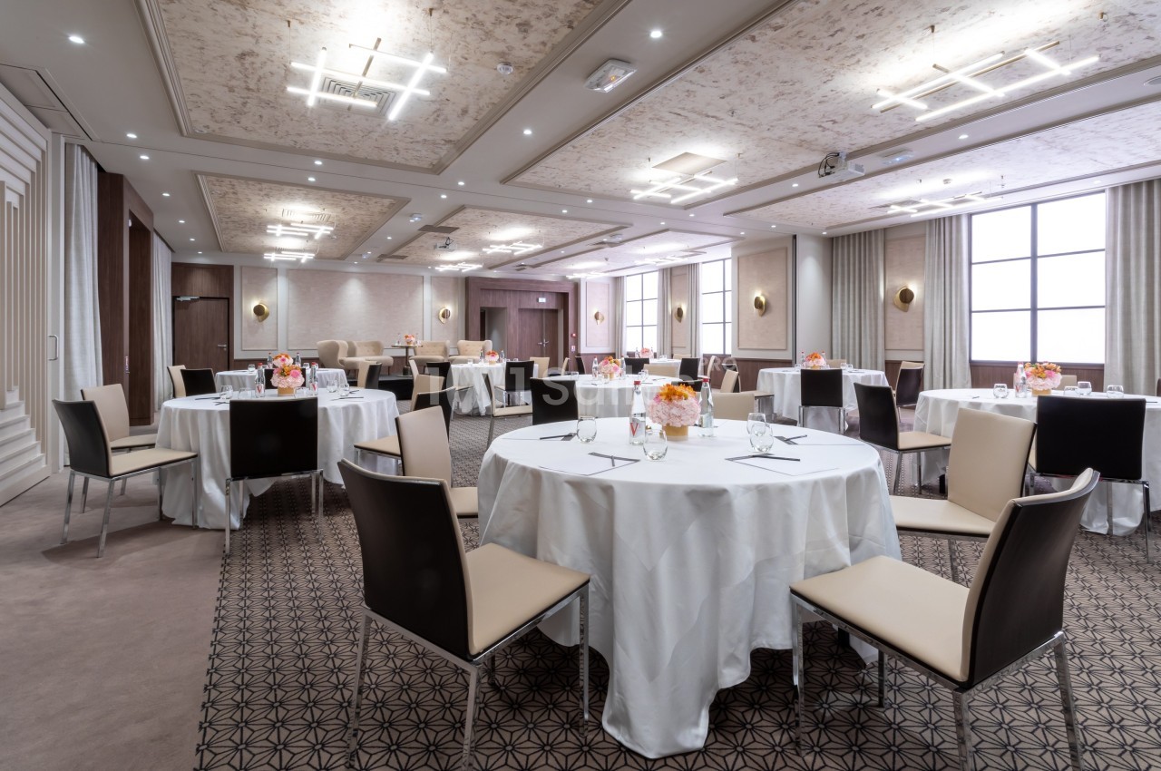 Salle de réception élégante avec tables rondes dressées, chaises modernes et décoration florale sur chaque table.