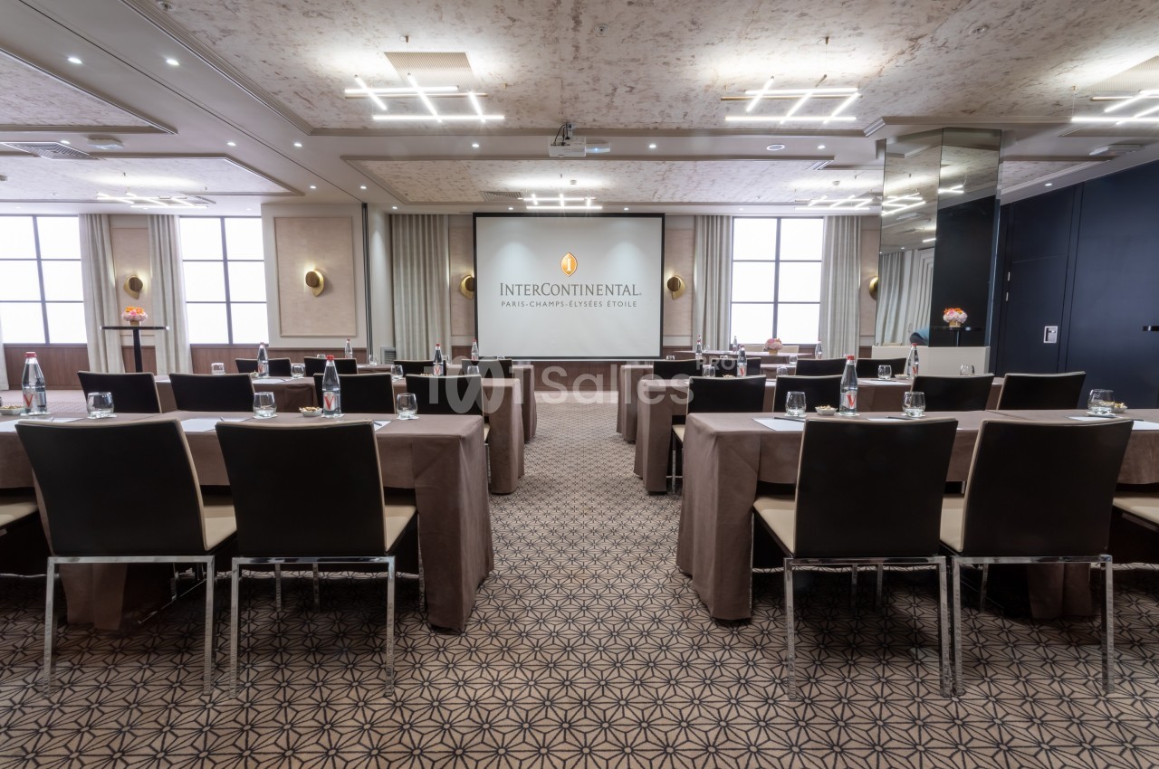 Salle de conférence lumineuse avec des rangées de tables et chaises, écran de projection et décor élégant.