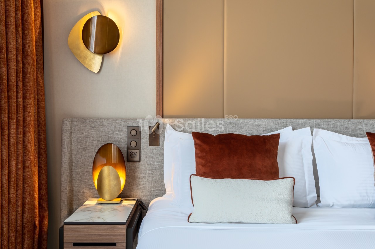Lit avec coussins blancs et rouille, table de chevet en marbre, lampe dorée et mur beige en arrière-plan.