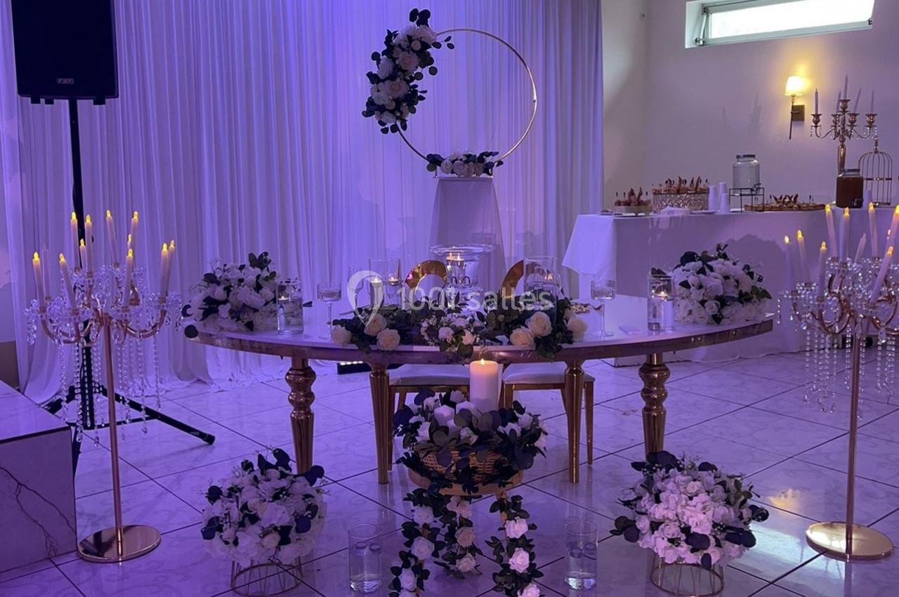 Location salle Brétigny-sur-Orge (Essonne) - La Joya Réception #8 Table décorée pour un événement avec fleurs blanches et violettes, chandeliers dorés et éclairage violet en arrière-plan.