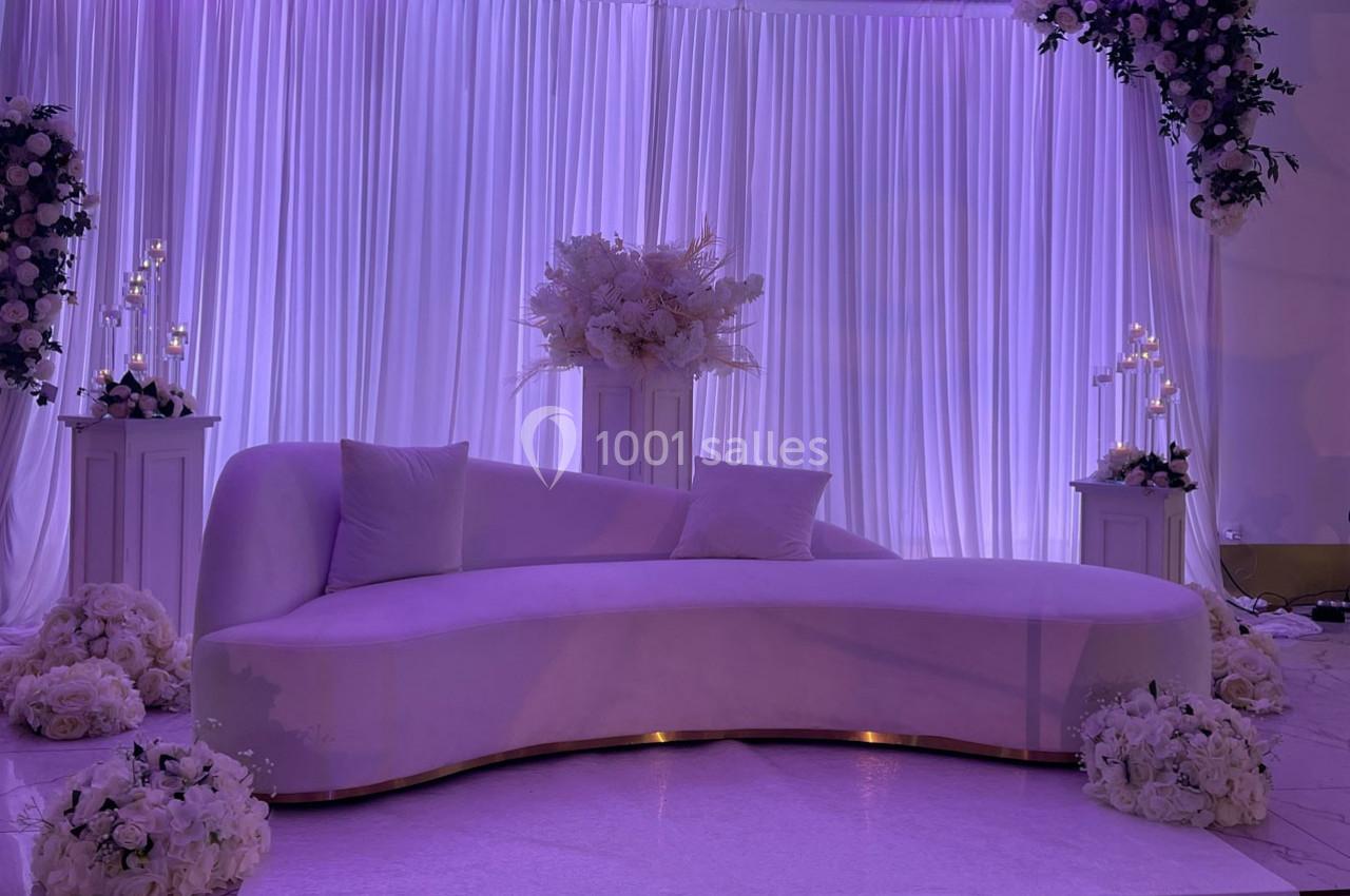 Location salle Brétigny-sur-Orge (Essonne) - La Joya Réception #4 Canapé courbé violet clair devant des rideaux plissés, entouré de fleurs blanches et de chandeliers décoratifs.