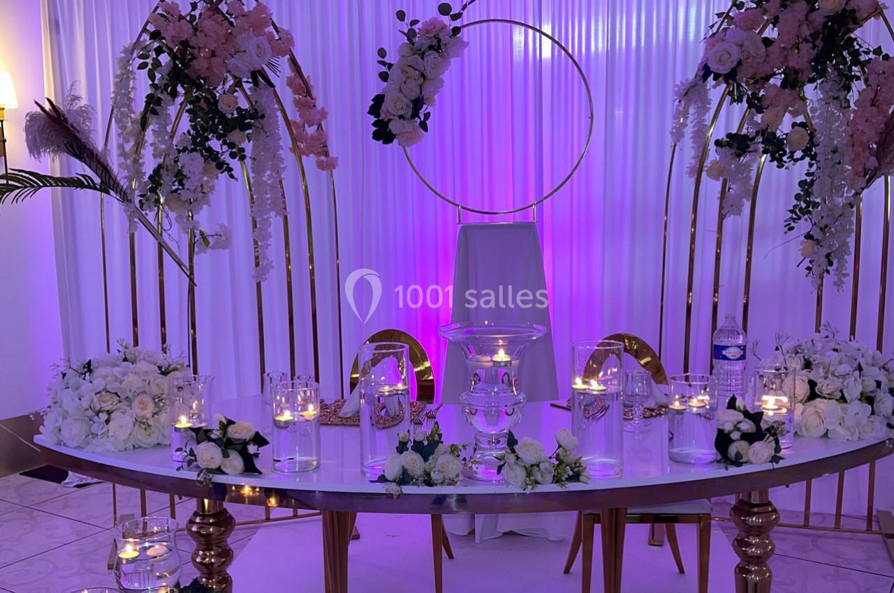 Location salle Brétigny-sur-Orge (Essonne) - La Joya Réception #14 Table décorée pour un événement avec fleurs blanches, bougies et arche dorée sur fond de rideaux blancs éclairés en violet.