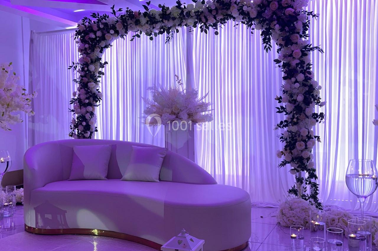 Location salle Brétigny-sur-Orge (Essonne) - La Joya Réception #16 Canapé blanc devant une arche florale ornée de roses et feuillages, éclairée par une lumière violette douce.