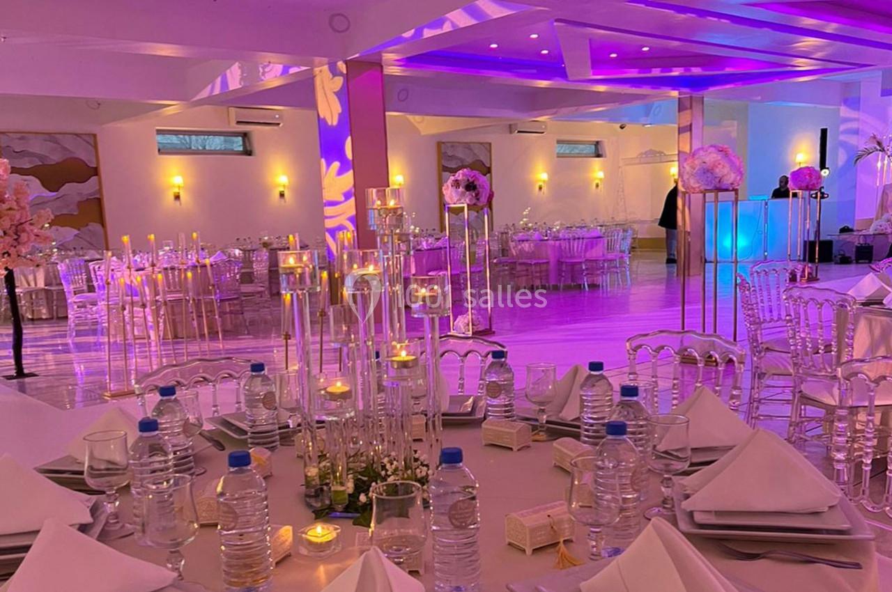 Salle de réception décorée avec des tables dressées, chandeliers, fleurs et éclairage violet.