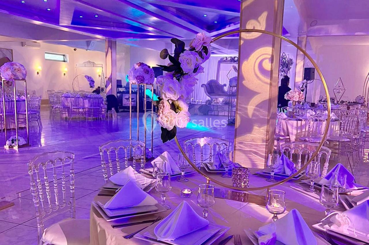 Location salle Brétigny-sur-Orge (Essonne) - La Joya Réception #6 Salle de réception élégante avec tables décorées de nappes blanches, chaises transparentes et éclairage violet.