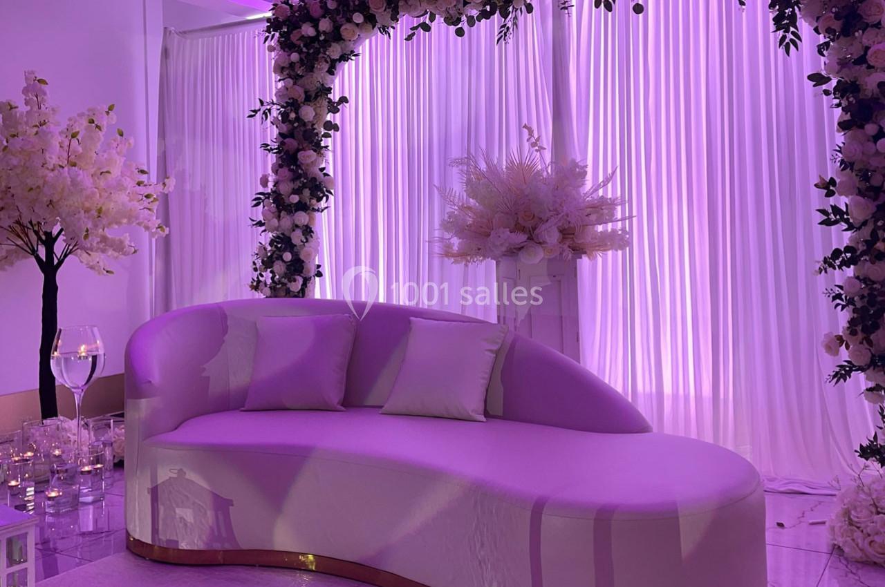 Location salle Brétigny-sur-Orge (Essonne) - La Joya Réception #19 Canapé courbé blanc devant des rideaux lumineux violets, décoré d'un arc floral et d'arrangements floraux élégants.