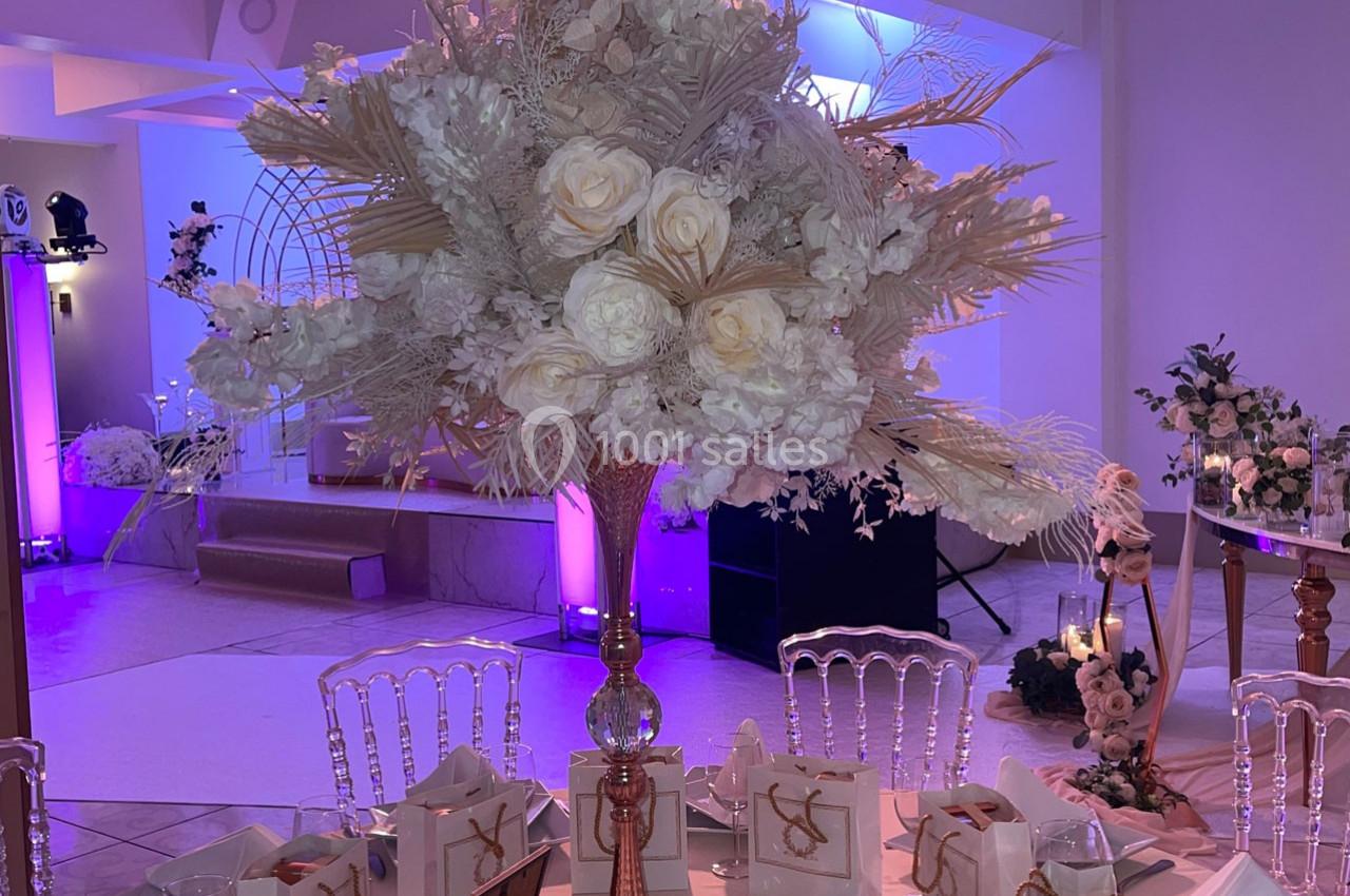 Location salle Brétigny-sur-Orge (Essonne) - La Joya Réception #13 Centre de table floral avec roses blanches et feuillage beige, dans une salle décorée de lumières violettes.