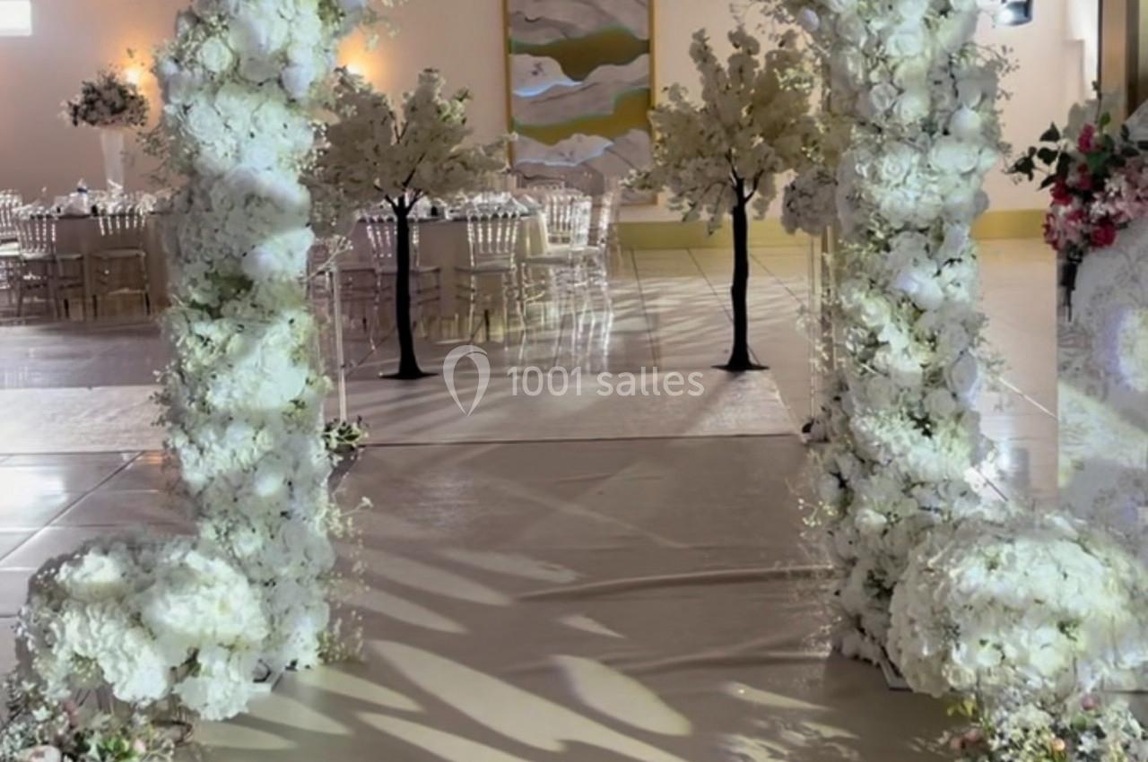 Location salle Brétigny-sur-Orge (Essonne) - La Joya Réception #7 Arche de mariage décorée de fleurs blanches, entourée de chaises transparentes et d'arbres artificiels.