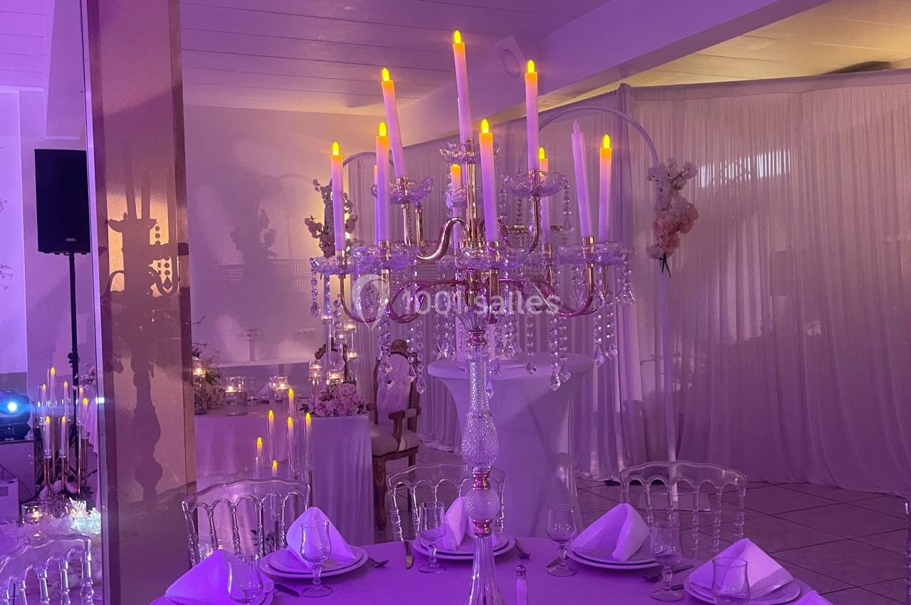 Location salle Brétigny-sur-Orge (Essonne) - La Joya Réception #9 Centre de table avec chandelier doré et bougies LED, entouré de tables dressées dans une salle éclairée en violet.