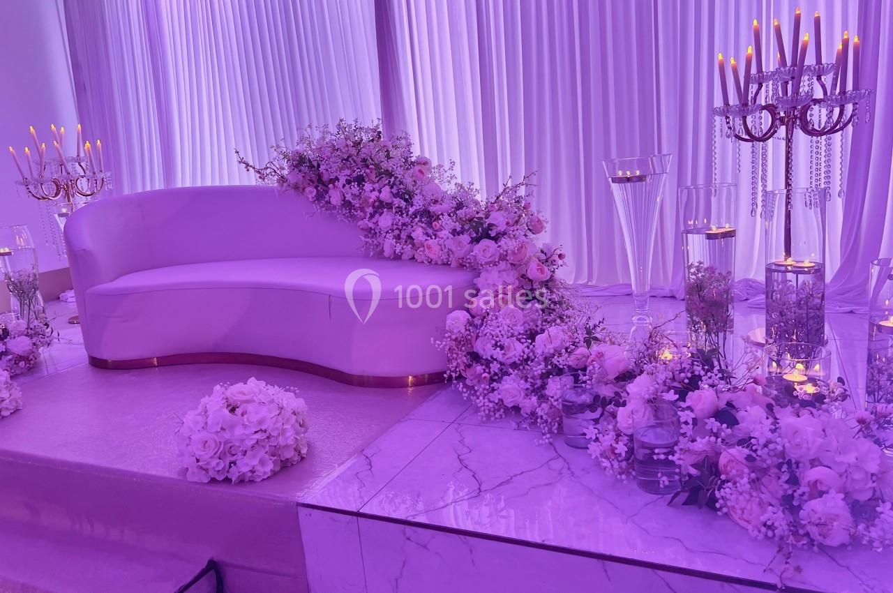 Location salle Brétigny-sur-Orge (Essonne) - La Joya Réception #3 Canapé blanc décoré de fleurs roses et blanches, entouré de chandeliers et de rideaux violets dans un décor élégant.