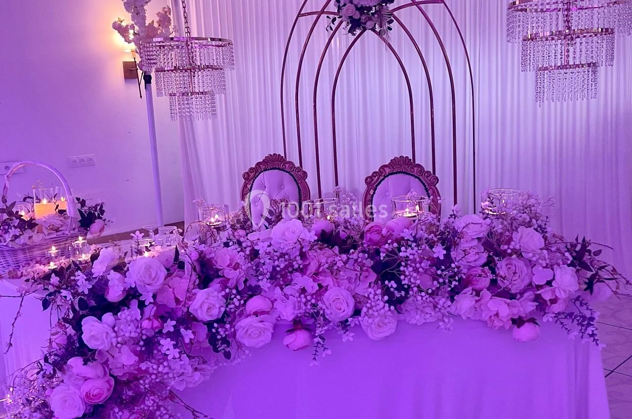 Location salle Brétigny-sur-Orge (Essonne) - La Joya Réception #5 Table ornée de fleurs roses et blanches, décorée pour un événement, avec arche métallique et éclairage tamisé.