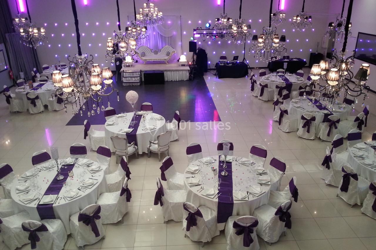 Salle de réception élégante avec tables rondes dressées, nappes blanches et décorations violettes sous des lustres suspendus.