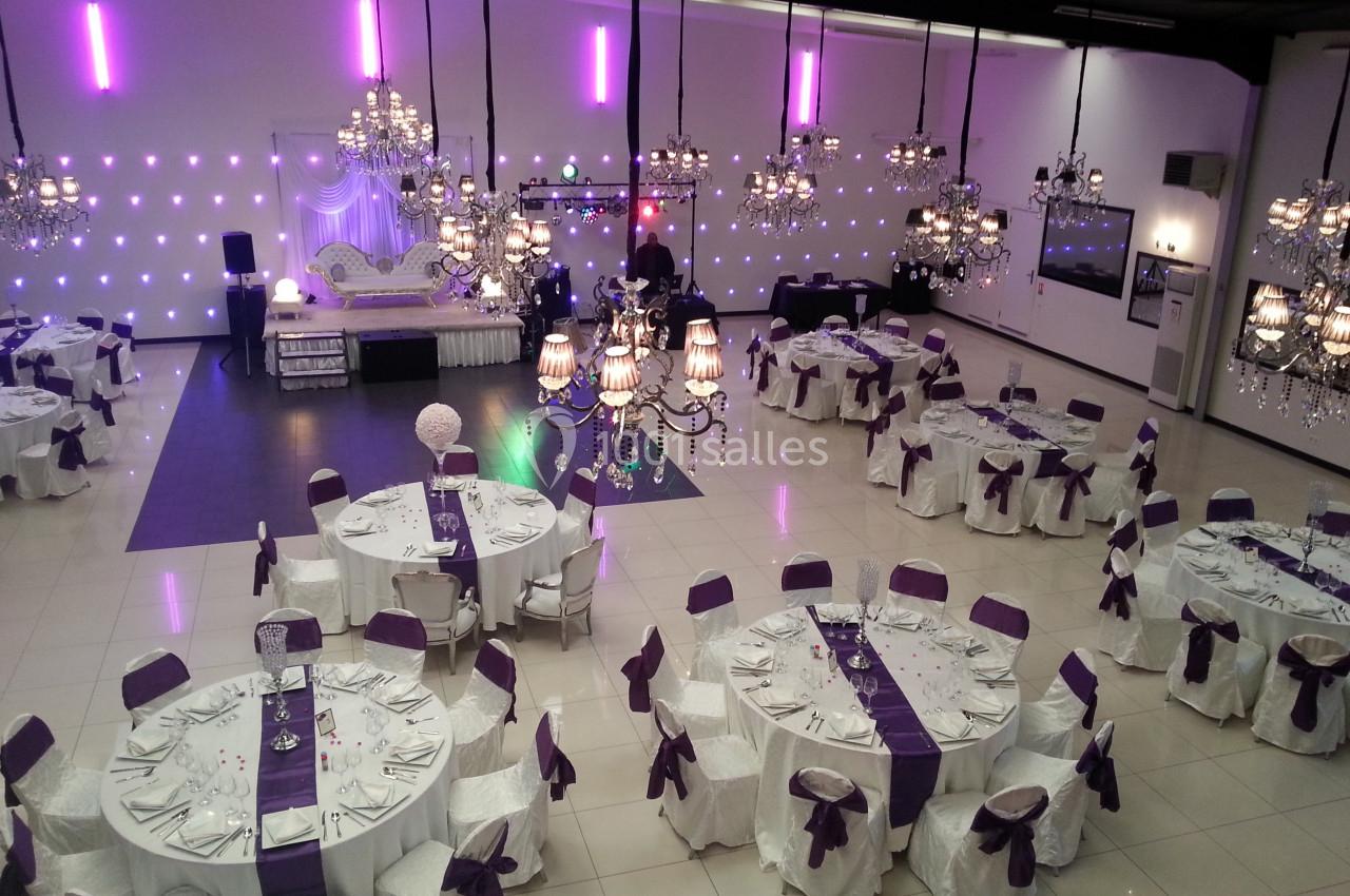 Salle de réception décorée avec des nappes blanches et des accents violets, éclairée par des lustres et des lumières murales.