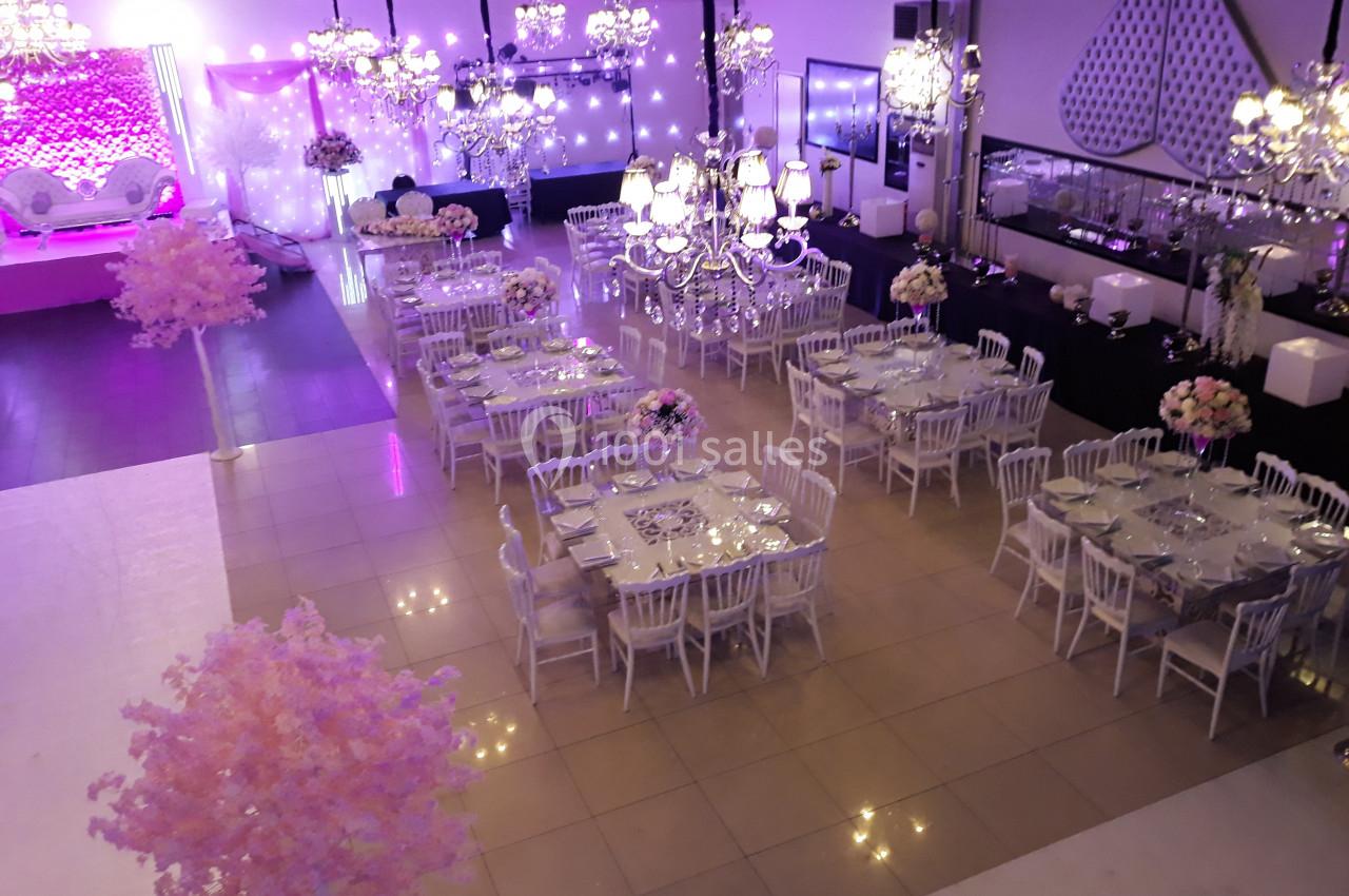 Salle de réception décorée avec des tables dressées, des chaises blanches, des lustres et des éclairages roses.