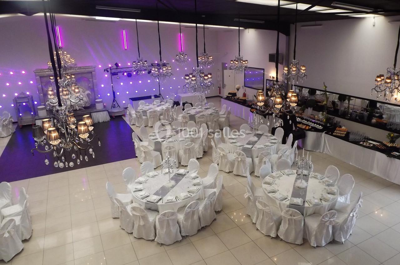 Salle de réception élégante avec tables rondes dressées, chaises blanches et lustres suspendus.