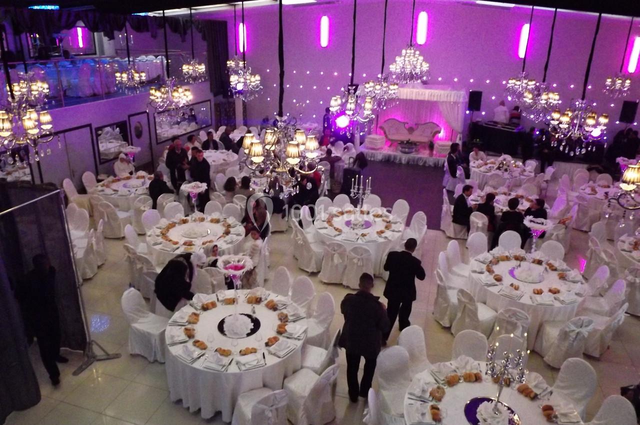 Salle de réception décorée avec des tables rondes dressées, des chaises blanches et des lustres suspendus.