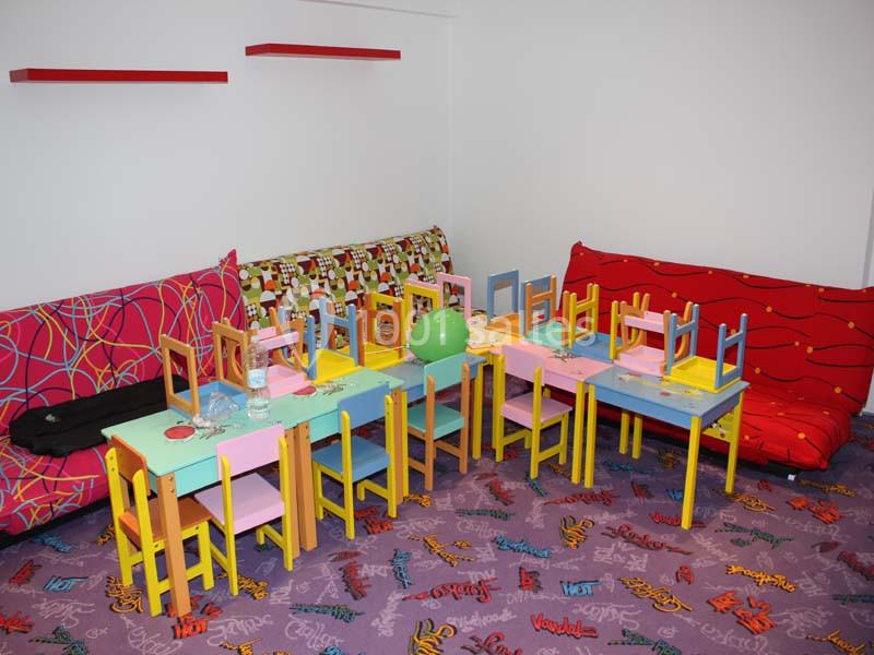 Salle colorée avec petites tables et chaises pour enfants, entourées de canapés aux motifs vifs sur un tapis décoré.