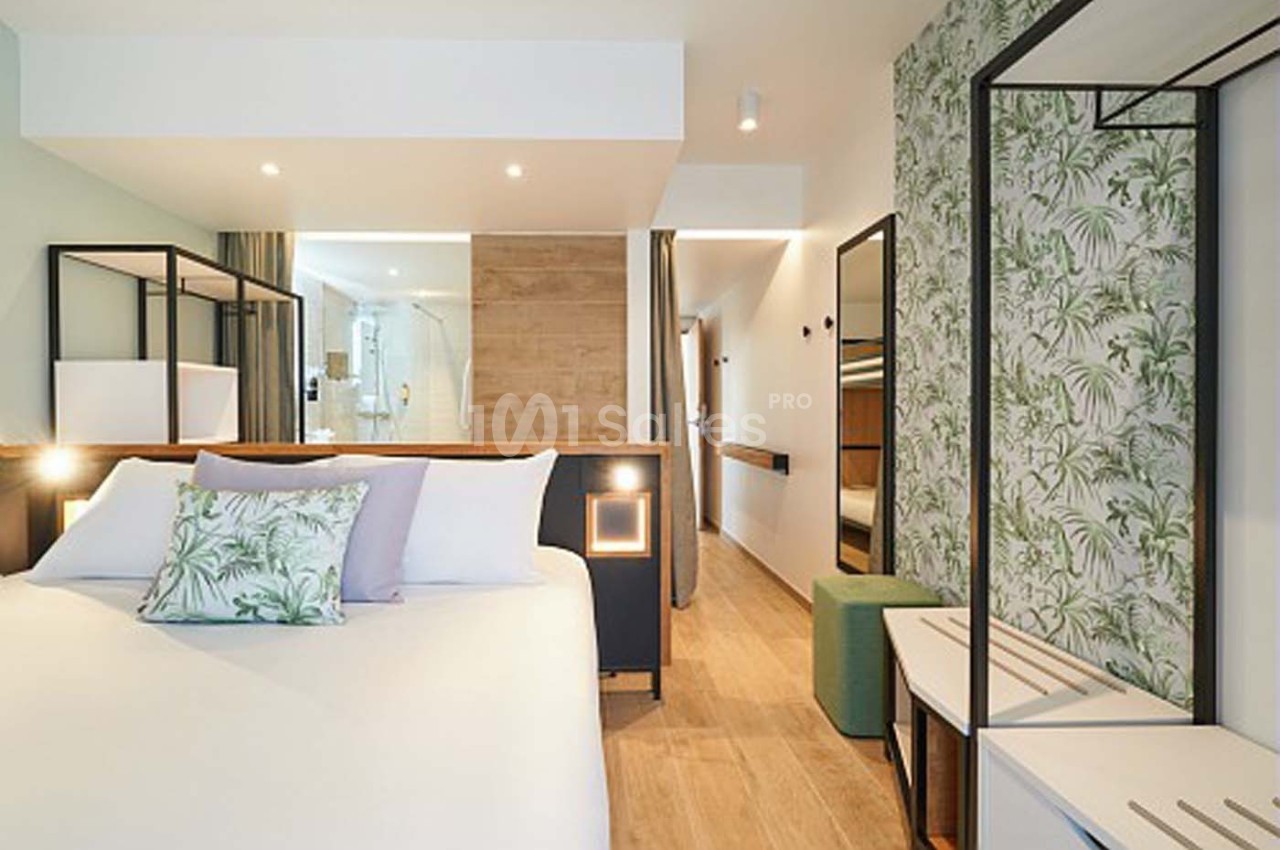 Chambre moderne avec lit double, décor végétal, éclairage doux et vue sur une salle de bain ouverte en arrière-plan.